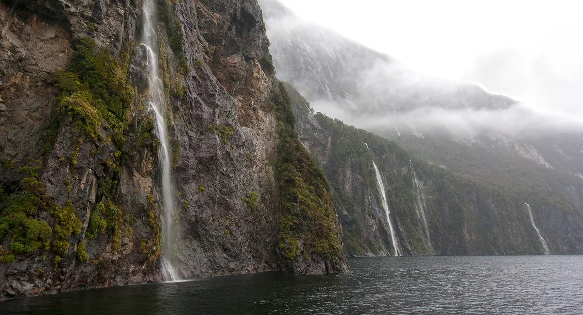 Waterfalls streaming down steep cliffs under low cloud after rain on a Milford Sound boat tour